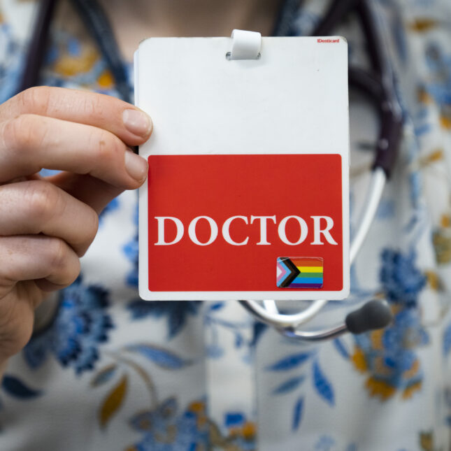 A newborn medicine doctor, holds up the backside of her name tag which reads "doctor" and has a pride flag sticker. -- health coverage from STAT